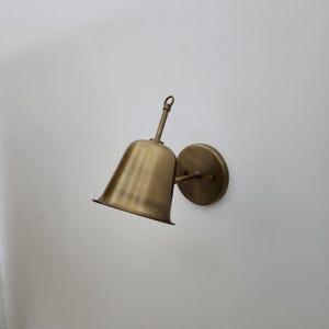 May include: A brass wall sconce featuring a bell-shaped shade and a circular mounting plate. The sconce has a vintage appearance, with a slightly aged finish. The shade is angled downwards. The fixture is designed for wall mounting.