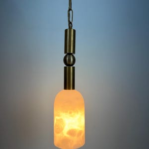 May include: A pendant light fixture with a warm, glowing, off-white, bottle-shaped shade. The fixture has brass-colored cylindrical and spherical accents, and a chain for hanging. The shade has a marbled, organic pattern.