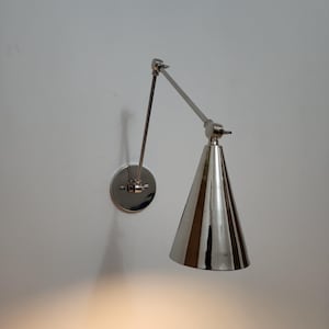 May include: A polished silver-tone wall-mounted lamp featuring a circular base plate, a slender adjustable arm with a central hinge, and a conical shade. The metal finish is reflective, and the design includes visible screws at the joints.