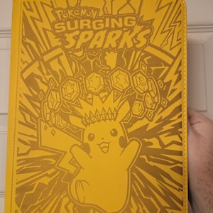 May include: Yellow card binder with gold Pikachu and "Pokemon Surging Sparks" text. The design includes a Pikachu graphic with a crown and lightning bolt pattern.