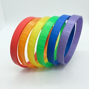 May include: A collection of seven colorful, rainbow-themed bracelets. The bracelets are arranged in a curved line, showcasing the colors red, orange, yellow, green, blue, and purple. Each bracelet has a textured surface.