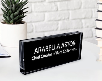 May include: A black desk nameplate with the name "ARABELLA ASTOR" and the title "Chief Curator of Rare Collections" in white lettering. The nameplate is on a white desk with a small potted plant and a stack of books in the background.