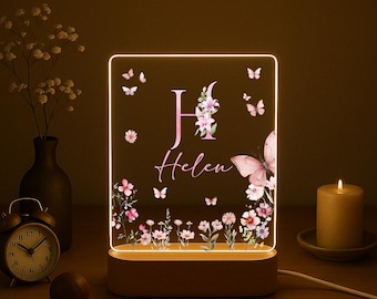 Pode incluir: Uma luz noturna personalizada com uma base de madeira. O painel de acrílico apresenta um design floral, borboletas e o nome "Helen" com a letra "H". A luz está acesa, criando um brilho quente. Uma vela e um despertador estão no fundo.