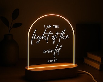 May include: A glowing LED night light with a wooden base. The clear acrylic panel displays the text "I AM THE light of the world" in a script font, with "JOHN 8:12" below. The light emits a warm, inviting glow.