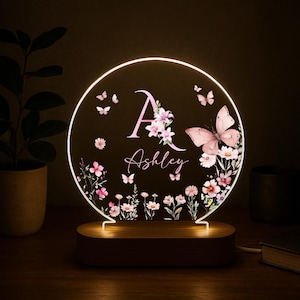 May include: A personalized night light with a circular design. The light features a pink butterfly and floral design, with the name "Ashley" and the initial "A" in pink. The light sits on a wooden base.