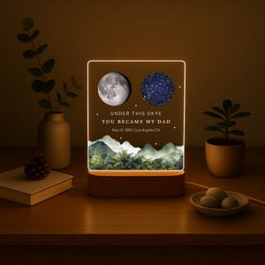 May include: A personalized night light with a brown base and a rectangular illuminated panel. The panel features a moon and a star map, with the text "UNDER THIS SKYE YOU BECAME MY DAD May 10, 1990 / Los Angeles CA". The design includes a mountain landscape.