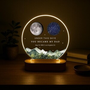 May include: A personalized night light with a circular design. It features a detailed moon and a star map, with the text "UNDER THIS SKYE YOU BECAME MY DAD" and a date. The base is wooden, and the light emits a warm glow.