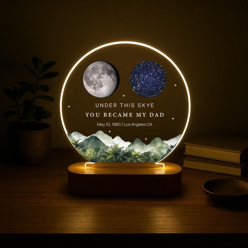 May include: A personalized night light with a circular design. It features a detailed moon and a star map, with the text "UNDER THIS SKYE YOU BECAME MY DAD" and a date. The base is wooden, and the light emits a warm glow.