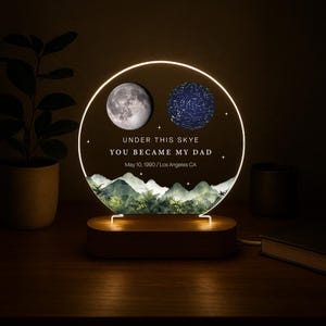 May include: A personalized night light with a wooden base. The circular light features a moon and a star map, with the text "UNDER THIS SKYE YOU BECAME MY DAD May 10, 1990 / Los Angeles CA". The light has a warm glow.