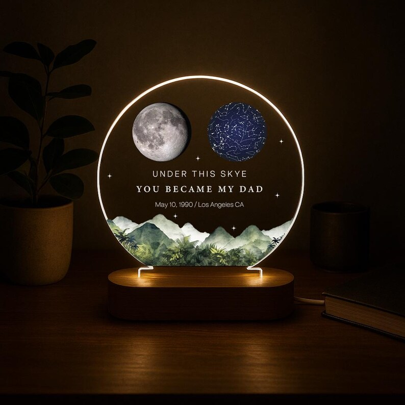 May include: A personalized night light with a wooden base. The circular light features a moon and a star map, with the text "UNDER THIS SKYE YOU BECAME MY DAD May 10, 1990 / Los Angeles CA". The light has a warm glow.