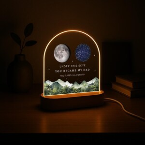 May include: A personalized night light with a wooden base and an arched acrylic panel. The panel features a moon and a star map, with the text "UNDER THIS SKYE YOU BECAME MY DAD" and a date. The light has a warm glow.