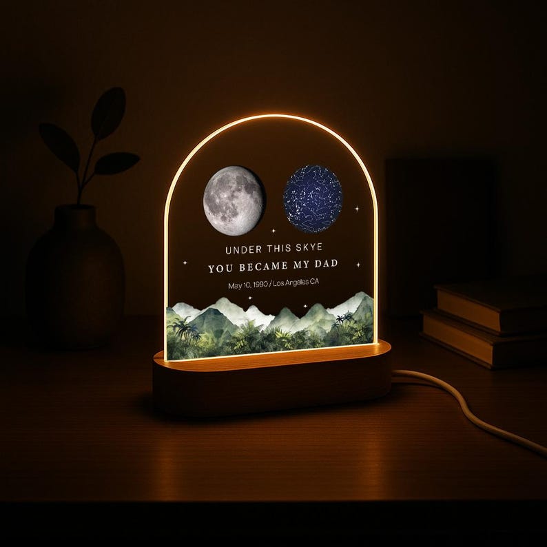 May include: A personalized night light with a wooden base and an arched acrylic panel. The panel features a moon and a star map, with the text "UNDER THIS SKYE YOU BECAME MY DAD" and a date. The light has a warm glow.