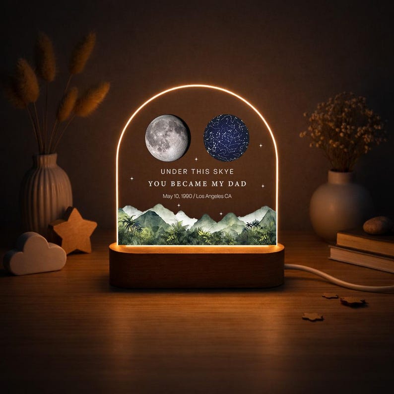 May include: A decorative night light with a wooden base and a clear acrylic panel. The panel features a moon and a star map, with the text "UNDER THIS SKYE YOU BECAME MY DAD May 10, 1900 / Los Angeles CA". The light has a warm glow.