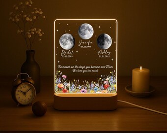 May include: A rectangular acrylic night light with a wooden base, featuring three moon phases, names, dates, and the text: The moons on the days you became our Mom. We love you so much. The design includes colorful wildflowers at the bottom.