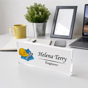 Puede incluir: Una placa de escritorio rectangular de acrílico transparente con un gráfico de un casco de seguridad amarillo y planos azules enrollados a la izquierda. El texto dice Helena Terry seguido del título Engineer en fuente negra.