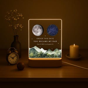 May include: A personalized night light with a wooden base. The acrylic panel features a moon and a star map, with the text "UNDER THIS SKYE YOU BECAME MY DAD" and the date May 10, 1990, Los Angeles CA. A candle and clock are also on the table.