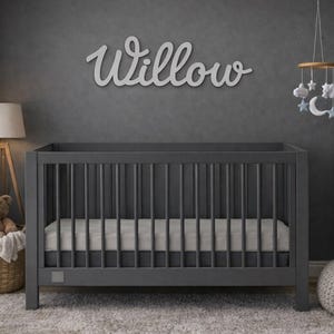 May include: A gray crib with a mattress is the focal point of a nursery. Above the crib, the name "Willow" is displayed in cursive. A mobile with clouds and stars hangs from the ceiling. A teddy bear sits in a wicker basket.