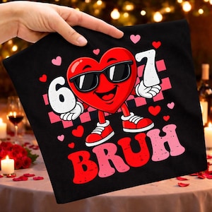 May include: Black t-shirt featuring a cartoon heart wearing sunglasses and sneakers. The heart holds the numbers 6 and 7, with the word "BRUH" below. Pink and red hearts and a checkered pattern complete the design.