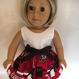 Puede incluir: Una muñeca con un top blanco sin mangas, una falda roja y negra con el logotipo de la Universidad de Alabama y zapatos negros. La falda tiene una cinta negra alrededor de la cintura.