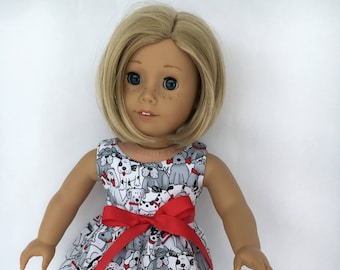 May include: A doll wearing a white dress with a red bow and a black and white dog print. The doll has blonde hair and blue eyes. The doll is wearing red shoes.