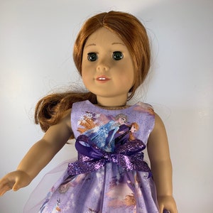 K&ouml;nnte beinhalten: Eine Puppe in einem lavendelfarbenen Kleid mit Frozen-Motiv. Das Kleid zeigt Bilder von Elsa und Anna, Schneeflocken und eine lila Glitzer-Schleife. Die Puppe hat rotes Haar und gr&uuml;ne Augen.