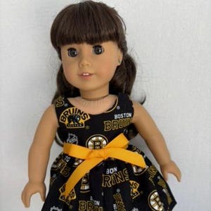 Puede incluir: Muñeca con un vestido negro con el logotipo y texto de los Boston Bruins en amarillo y blanco, con un lazo amarillo en la cintura. La muñeca tiene el pelo castaño y lleva zapatos negros. El vestido tiene el texto "BOSTON BRUINS".