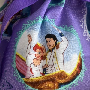 May include: Purple fabric bag with a repeating pattern of Ariel and Prince Eric from The Little Mermaid. The pattern features a light blue background with white and green accents. The bag has a purple ribbon tie.