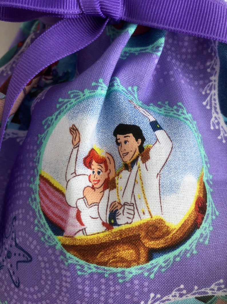 May include: Purple fabric bag with a repeating pattern of Ariel and Prince Eric from The Little Mermaid. The pattern features a light blue background with white and green accents. The bag has a purple ribbon tie.