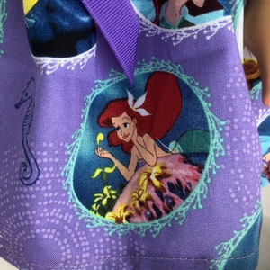 May include: Purple fabric with a repeating pattern of Ariel, the Little Mermaid, in different scenes. The fabric has a light blue, white, and green border around each image. The fabric is sewn with a purple ribbon.