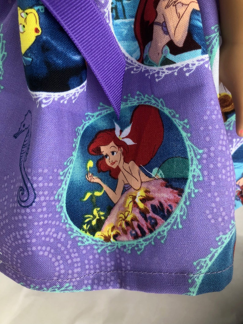 May include: Purple fabric with a repeating pattern of Ariel, the Little Mermaid, in different scenes. The fabric has a light blue, white, and green border around each image. The fabric is sewn with a purple ribbon.