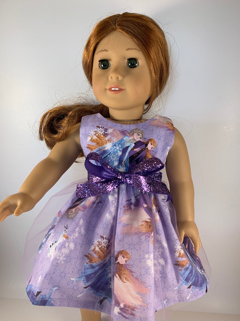 K&ouml;nnte beinhalten: Eine Puppe in einem lavendelfarbenen Kleid mit Frozen-Motiv. Das Kleid zeigt Bilder von Elsa und Anna, Schneeflocken und eine lila Glitzer-Schleife. Die Puppe hat rotes Haar und gr&uuml;ne Augen.
