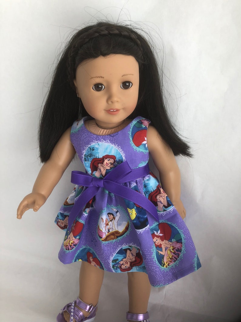 May include: A purple dress with a print of Ariel from The Little Mermaid. The dress has a purple ribbon tied around the waist. The doll is wearing the dress and has long black hair.