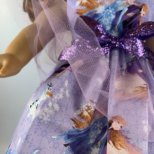 K&ouml;nnte beinhalten: Ein lavendelfarbenes Puppenkleid mit Figuren aus dem Film Frozen. Das Kleid hat ein gedrucktes Muster von Elsa, Anna und Olaf, mit einer passenden Schleife und T&uuml;llakzenten. Das Kleid ist f&uuml;r eine Puppe entworfen.