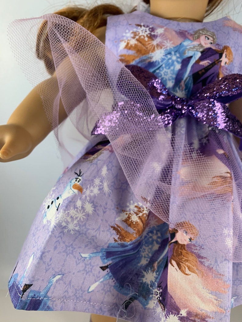 K&ouml;nnte beinhalten: Ein lavendelfarbenes Puppenkleid mit Figuren aus dem Film Frozen. Das Kleid hat ein gedrucktes Muster von Elsa, Anna und Olaf, mit einer passenden Schleife und T&uuml;llakzenten. Das Kleid ist f&uuml;r eine Puppe entworfen.