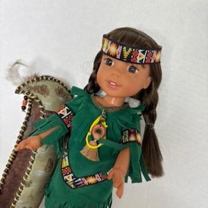 May include: A doll dressed in a Native American costume. The doll wears a green suede-like dress with fringe, a patterned headband, and matching boots. A decorative carrier is behind the doll.