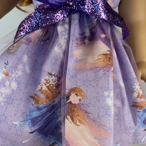K&ouml;nnte beinhalten: Ein Puppenkleid mit einem lila-wei&szlig;en Charakterdruck, einem T&uuml;ll-Overlay und einer glitzernden lila Schleife. Das Kleid ist f&uuml;r eine Puppe entworfen.