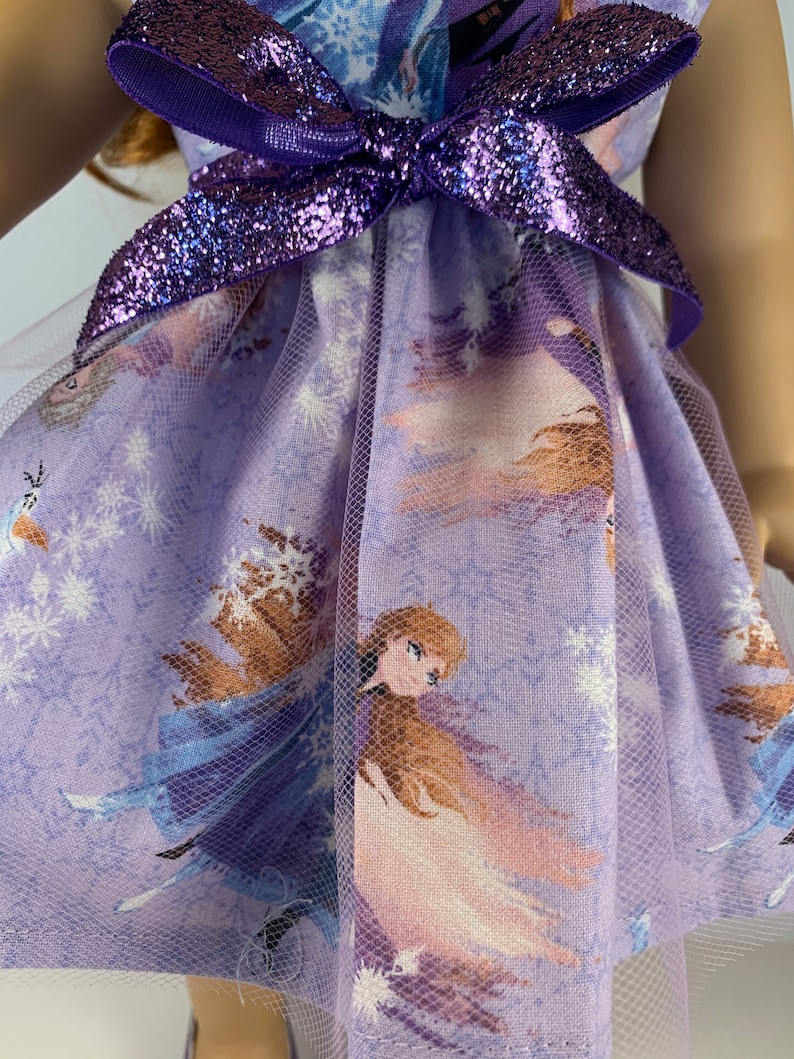 K&ouml;nnte beinhalten: Ein Puppenkleid mit einem lila-wei&szlig;en Charakterdruck, einem T&uuml;ll-Overlay und einer glitzernden lila Schleife. Das Kleid ist f&uuml;r eine Puppe entworfen.
