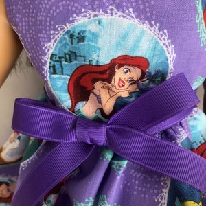 May include: Purple dress with a print of Ariel from The Little Mermaid. The dress has a purple bow at the waist.