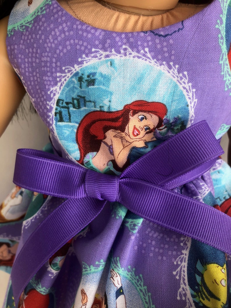 May include: Purple dress with a print of Ariel from The Little Mermaid. The dress has a purple bow at the waist.