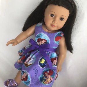 May include: A purple dress with a print of Ariel from The Little Mermaid. The dress has a purple ribbon tied around the waist. The doll is wearing the dress and has long black hair.