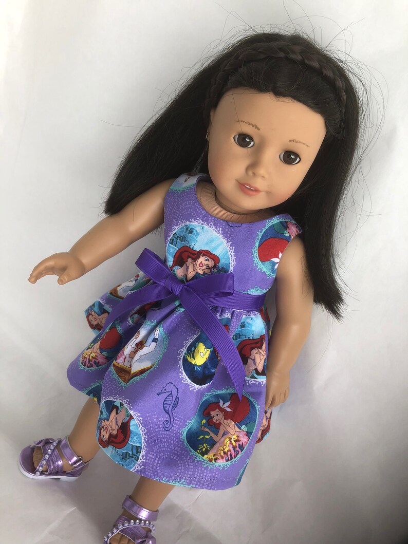 May include: A purple dress with a print of Ariel from The Little Mermaid. The dress has a purple ribbon tied around the waist. The doll is wearing the dress and has long black hair.