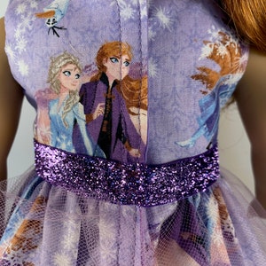K&ouml;nnte beinhalten: Ein Puppenkleid mit Frozen-Motiv. Das lavendelfarbene Oberteil zeigt Elsa, Anna und Olaf, mit einem glitzernden lila G&uuml;rtel. Der Rock ist lavendelfarben mit einem transparenten Overlay.