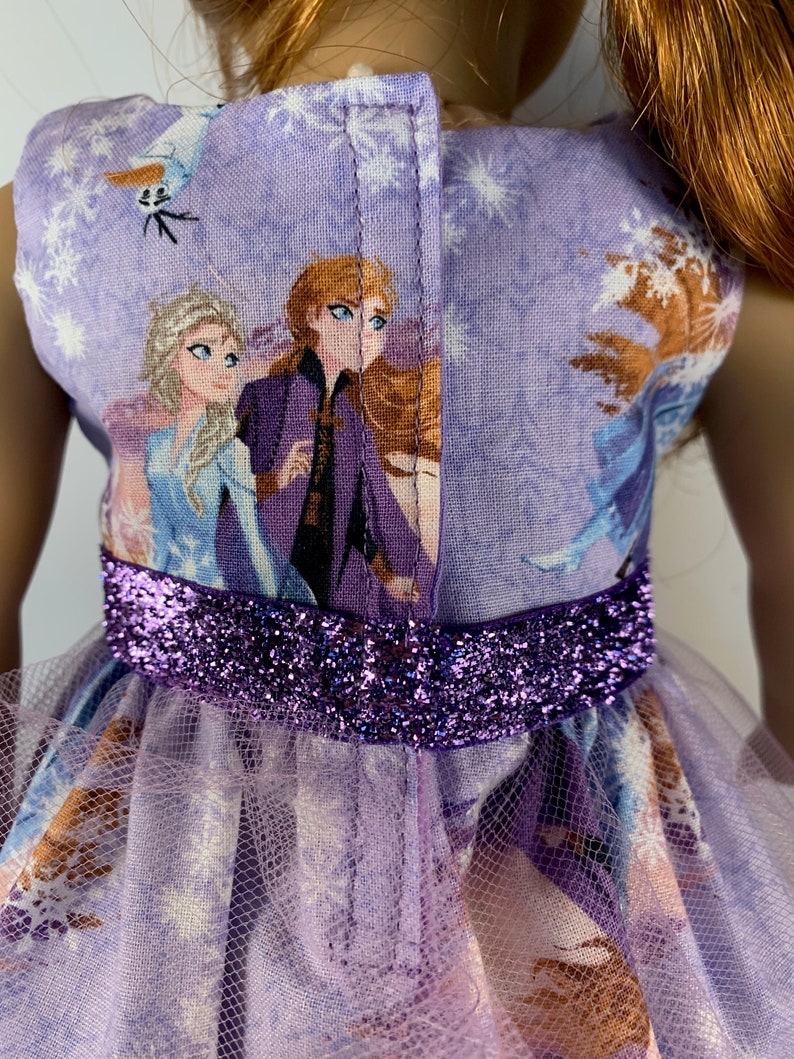 K&ouml;nnte beinhalten: Ein Puppenkleid mit Frozen-Motiv. Das lavendelfarbene Oberteil zeigt Elsa, Anna und Olaf, mit einem glitzernden lila G&uuml;rtel. Der Rock ist lavendelfarben mit einem transparenten Overlay.