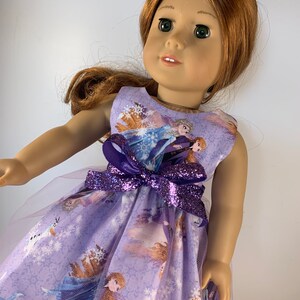 K&ouml;nnte beinhalten: Eine Puppe in einem &auml;rmellosen, lavendelfarbenen Kleid mit einem Aufdruck von Figuren aus dem Film "Frozen". Das Kleid hat eine lila Glitzer-Schleife und einen T&uuml;llrock. Die Puppe hat rotes Haar und gr&uuml;ne Augen.