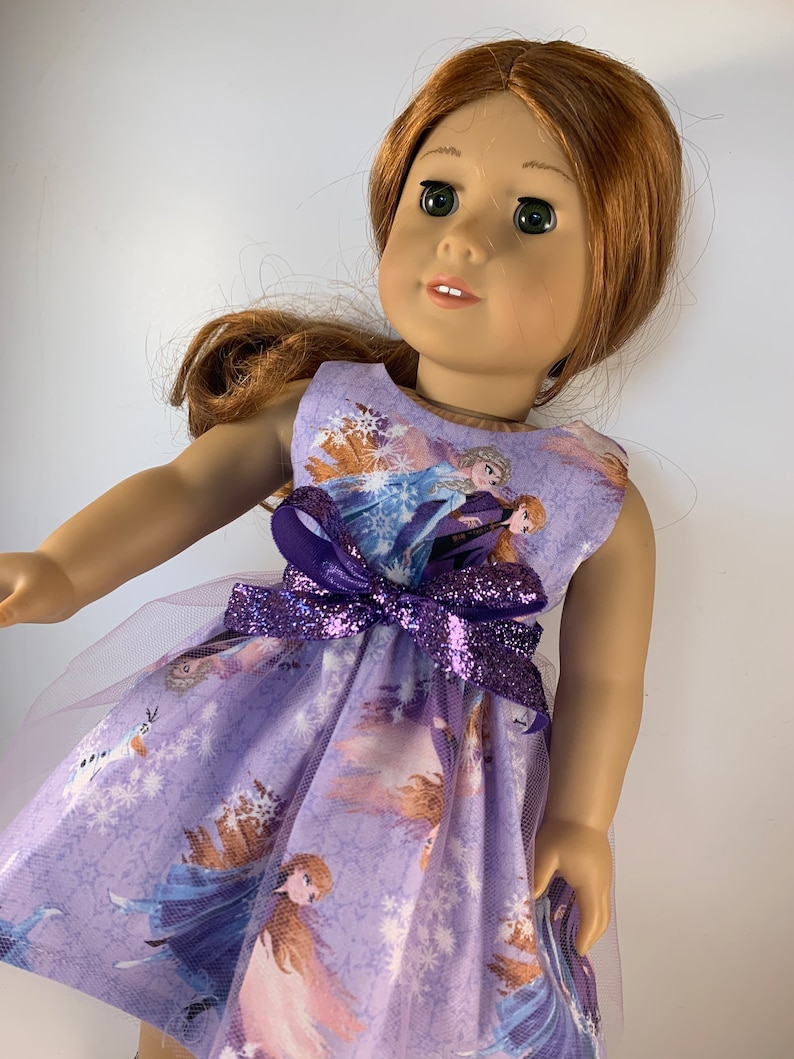K&ouml;nnte beinhalten: Eine Puppe in einem &auml;rmellosen, lavendelfarbenen Kleid mit einem Aufdruck von Figuren aus dem Film "Frozen". Das Kleid hat eine lila Glitzer-Schleife und einen T&uuml;llrock. Die Puppe hat rotes Haar und gr&uuml;ne Augen.