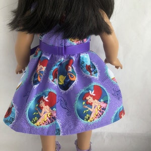 May include: A purple dress with a  pattern of Ariel from The Little Mermaid. The dress has a purple ribbon around the waist and is worn by a doll.