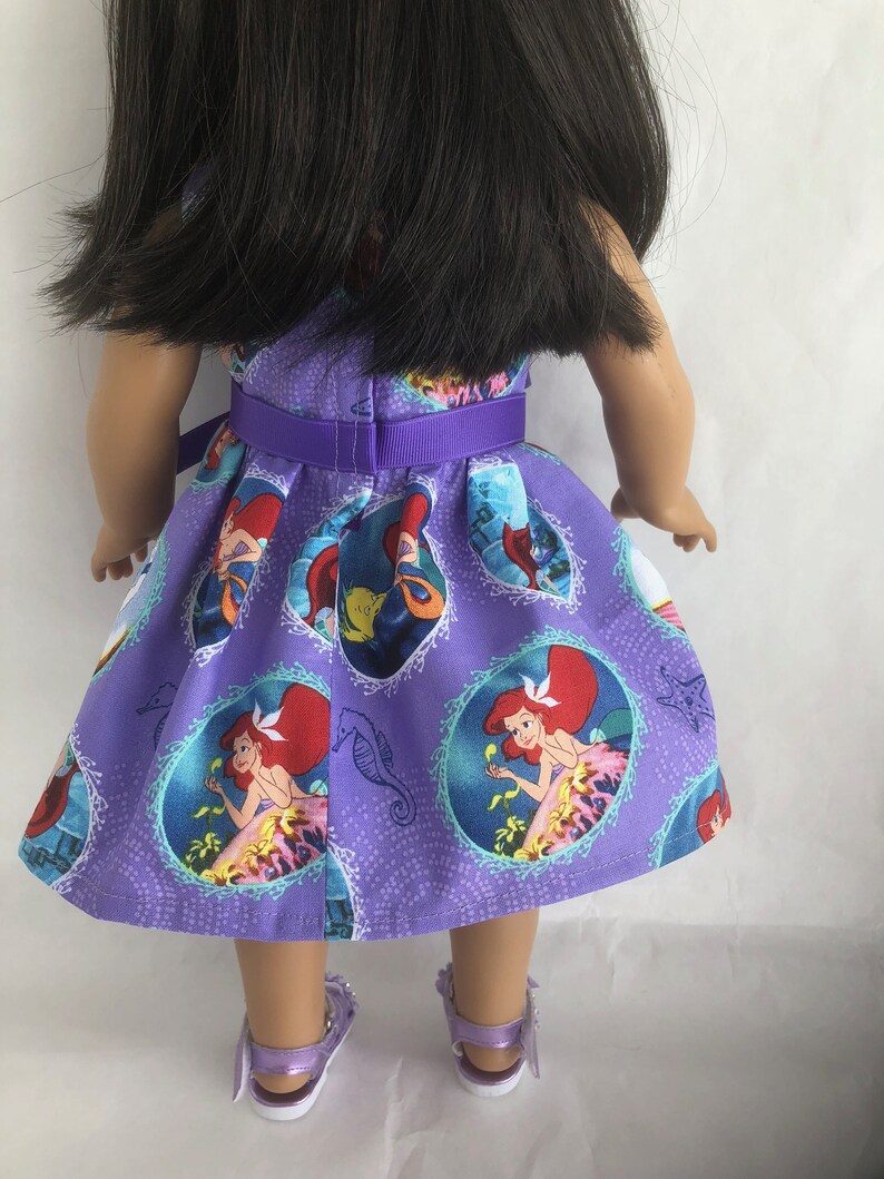 May include: A purple dress with a  pattern of Ariel from The Little Mermaid. The dress has a purple ribbon around the waist and is worn by a doll.