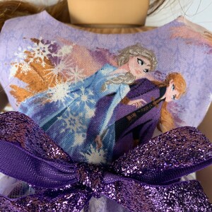K&ouml;nnte beinhalten: Ein Puppenkleid mit einem lavendelfarbenen Oberteil mit einem Aufdruck von zwei animierten Figuren in Blau und Lila. Eine gro&szlig;e, glitzernde lila Schleife ziert die Taille. Das Kleid hat einen T&uuml;llrock.