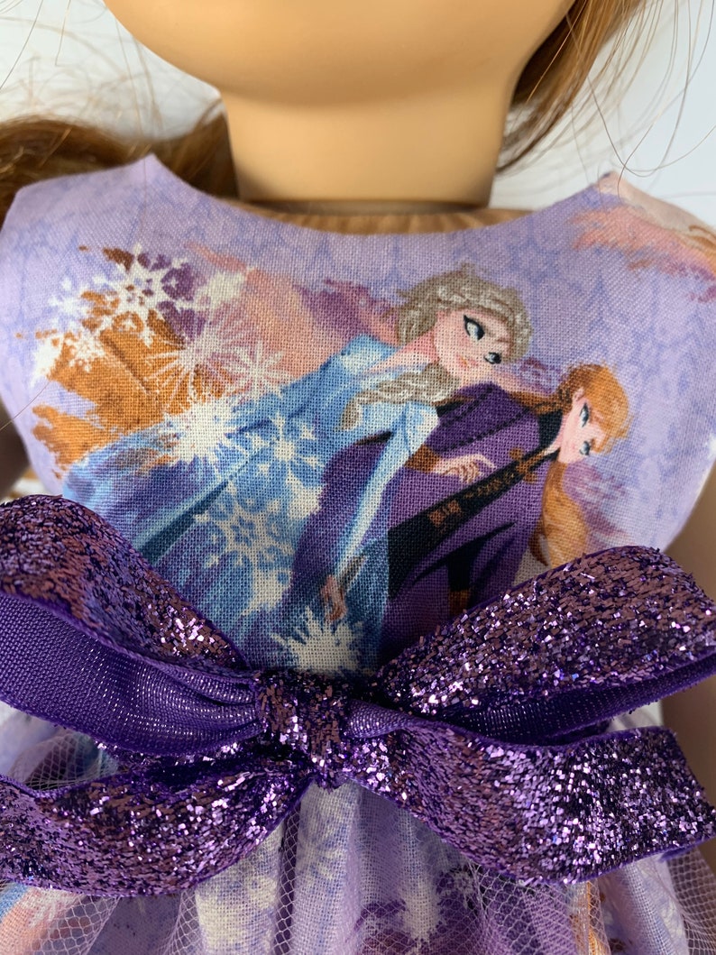 K&ouml;nnte beinhalten: Ein Puppenkleid mit einem lavendelfarbenen Oberteil mit einem Aufdruck von zwei animierten Figuren in Blau und Lila. Eine gro&szlig;e, glitzernde lila Schleife ziert die Taille. Das Kleid hat einen T&uuml;llrock.
