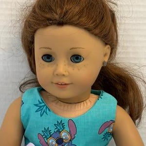 Puede incluir: Muñeca con ojos azules, pecas y cabello castaño rojizo recogido en una cola de caballo. La muñeca lleva un vestido turquesa con un estampado de Stitch y un lazo lavanda. La muñeca tiene un tono de piel claro.